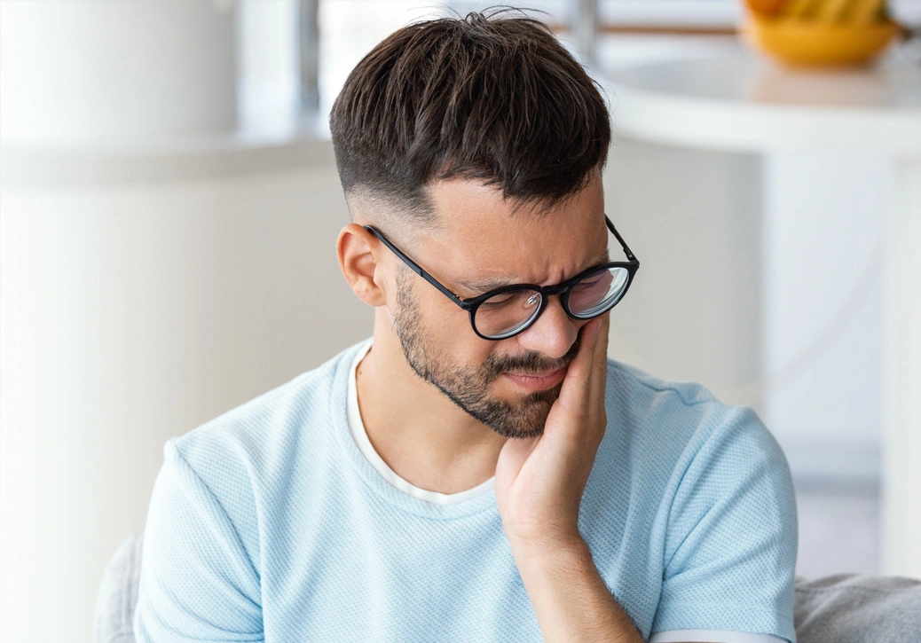 Man wearing glasses and light blue shirt, resting chin on hand, looking thoughtful in bright indoor setting.